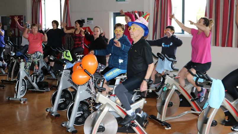 A man in a black top, wearing a pink hat with candles on it rides an Exercycle with orange balloons on the front while a group of people around him Excercycles wave their hands in the air