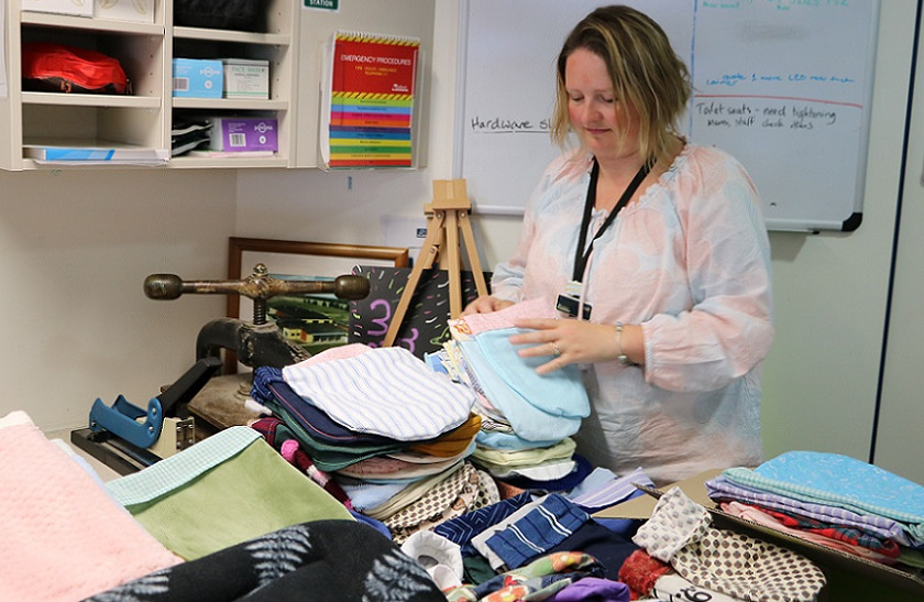 Woman lifts a pair of blue and pink sewn large pocket shaped items from the top of a table covered in similar multi coloured stacked items
