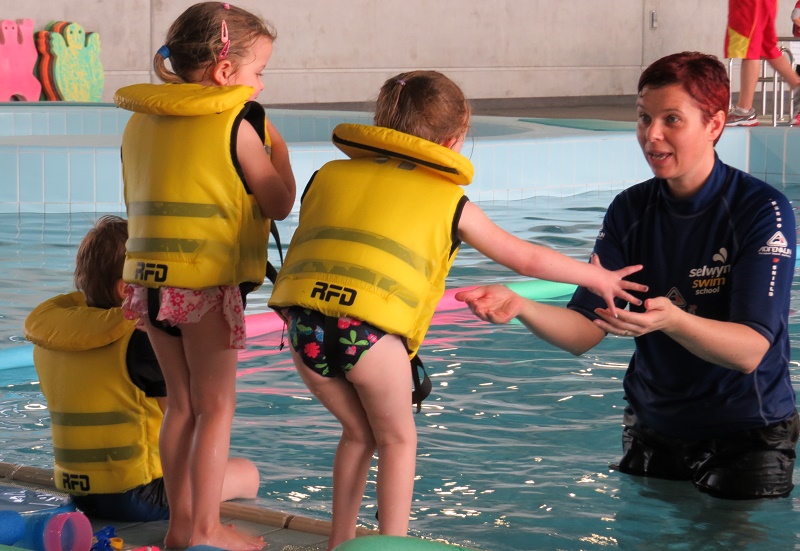 A woman with short red hair in a dark blue swimming top stands in a pool holding her arms out towards three children in lifejackets standing on the edge of the pool