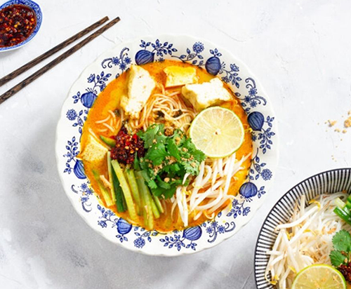 Malaysian noodles orange soup with lemon, cucumber, bean sprouts in a china blue and white bowl with chopsticks on the side and a chilli dip 