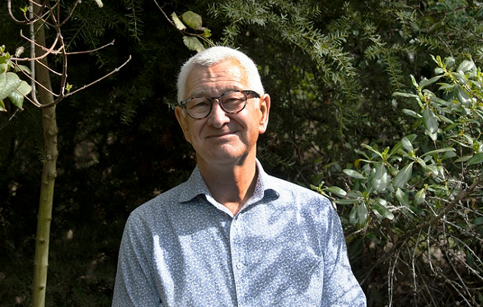 A man with short white hair and glasses stands in front of some bush
