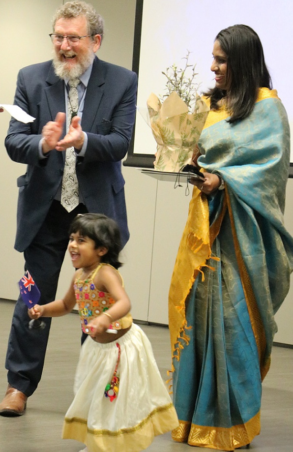 A woman in a sari, carrying a plant and Deputy Mayor Malcolm Lyall laugh as a young girl in a sari runs away smiling carrying a small New Zealand flag