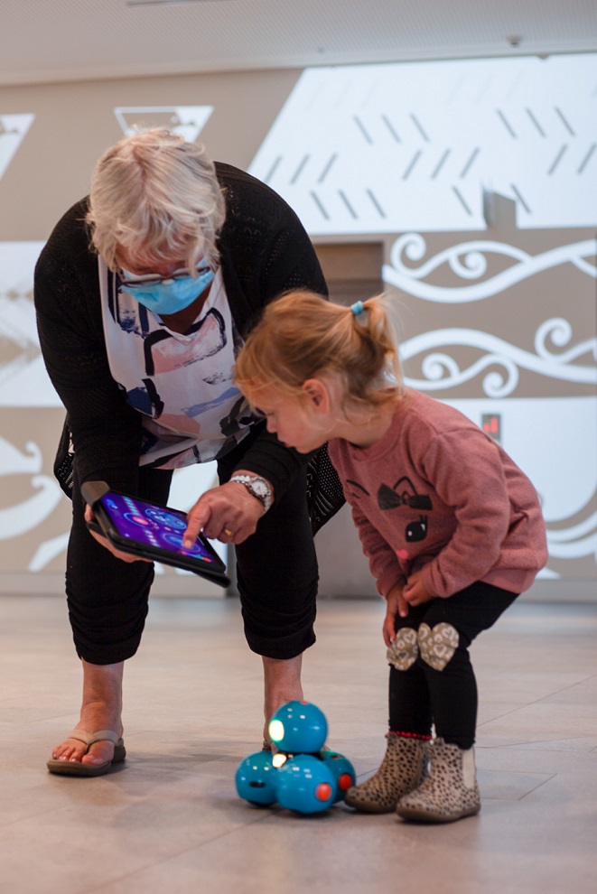 A grandmother and granddaughter look at the ipad controls for a small blue robot they are driving across the ground floor at Te Ara Ātea