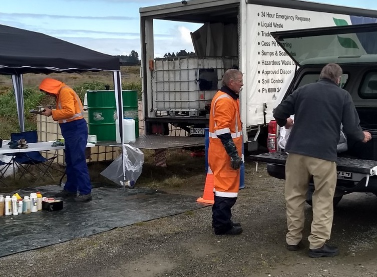 A man with his back to the camera unloads something from the back of a pick up while a man in orange high visibility overalls waits to receive it and another man in high visibility orange overalls inspects items in a marquee