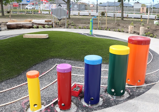 Colourful steel tubes in front of an astroturfed mound with an outdoor xylophone and log seat in the background