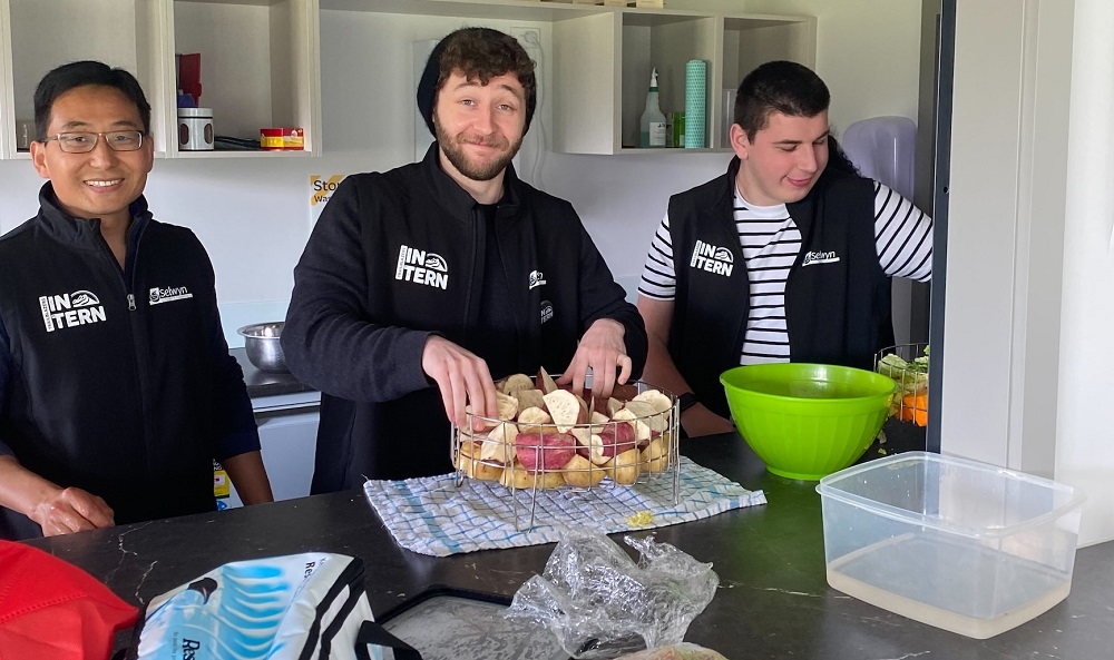 Three men in black jerseys labelled intern, in a kitchen mixing some food ready to cook