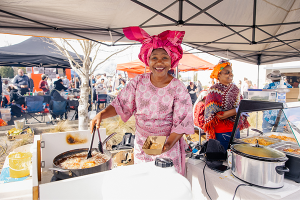 Nigerian lady in pink at food stall holding tongs stirring a dish in a pot 