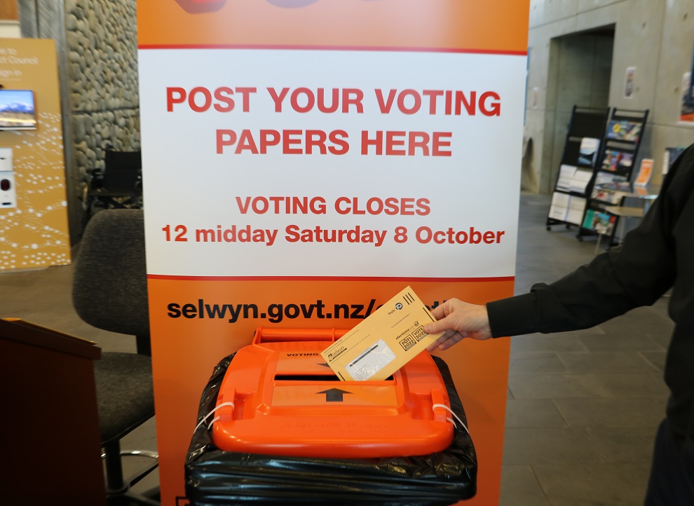 An arm posting a voting paper into a ballot box underneath a sign saying post your voting papers here