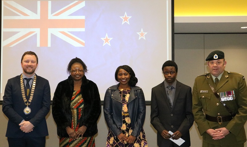 Five people, Mayor Sam Broughton, Dorothy Chilwalo, Angela Yendama, Chinjila Lunda and Major Grant Payton in army uniform stand in a line in front of a screen with the New Zealand flag projected on it