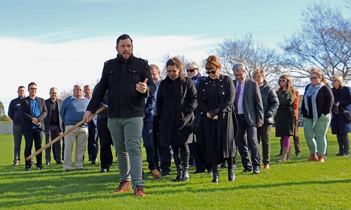 A young maori man with a beard and a black jacket gestures with a tokotoko as he leads a group walking with bowed heads across a park