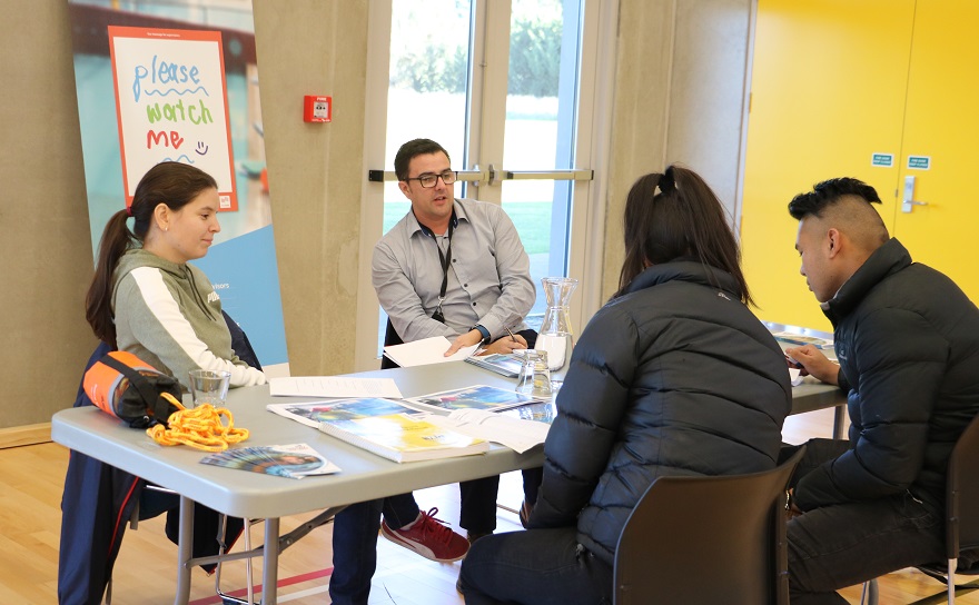Two students in black puffer jackets and black hair sit with backs to camera reading something on a grey table opposite a woman and a man