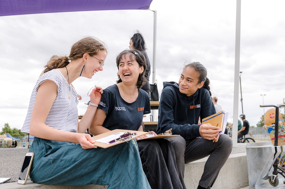 Three youth councillors, two in black BOOST Selwyn Youth Council t-shirts, laughing together while having a discussion sitting next to a skate park