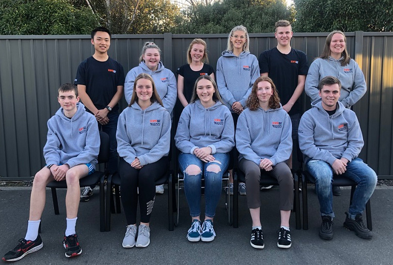 11 teenagers in two rows, the back row standing the front row sitting all wearing matching grey hoodies or black t-shirts
