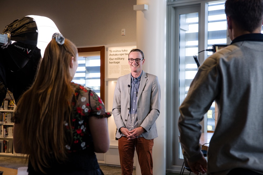 Image shows the back of an interviewer, camera man and a large stand lamp with a smiling man in a suit facing them looking towards the interviewer
