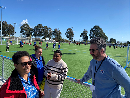 Three woman and a man stand in front of a soccer field with people on the field in the backgroun