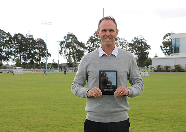 Council male employee stands with award plaque in hand with Foster Park greenery in background and kids playing