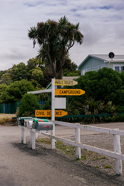 Sign post pointing to Campground and Civil Defence outside letter box and in front of cabbage tree and house