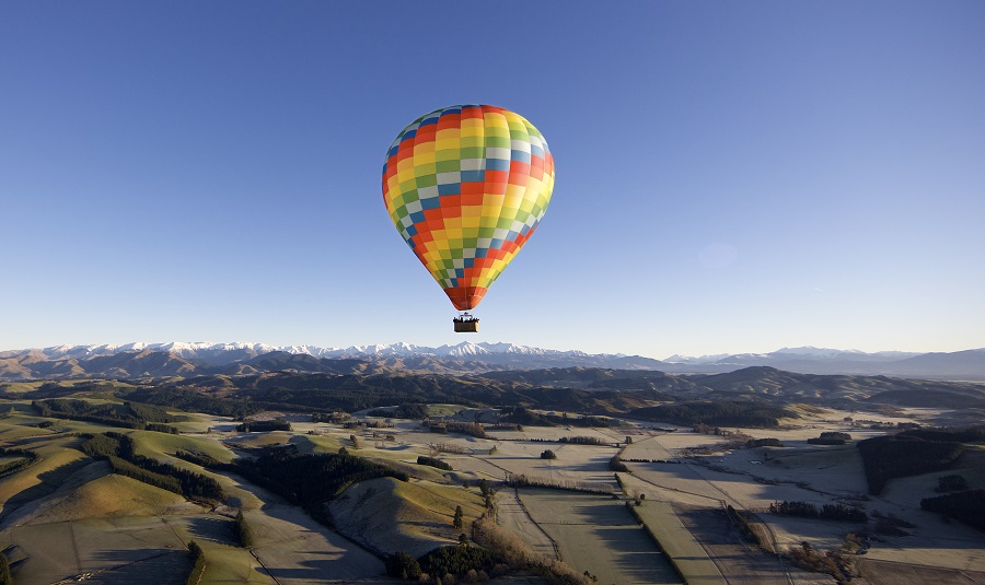 A hot air balloon in rainbow colours floating in a blue sky above the Canterbury Plains