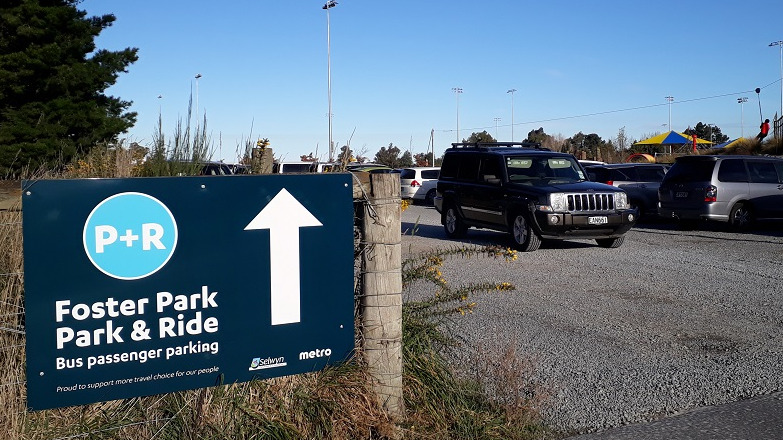 Picture of a blue sign on a fence saying park and ride, with cars parked in the background and black car driving out the gate
