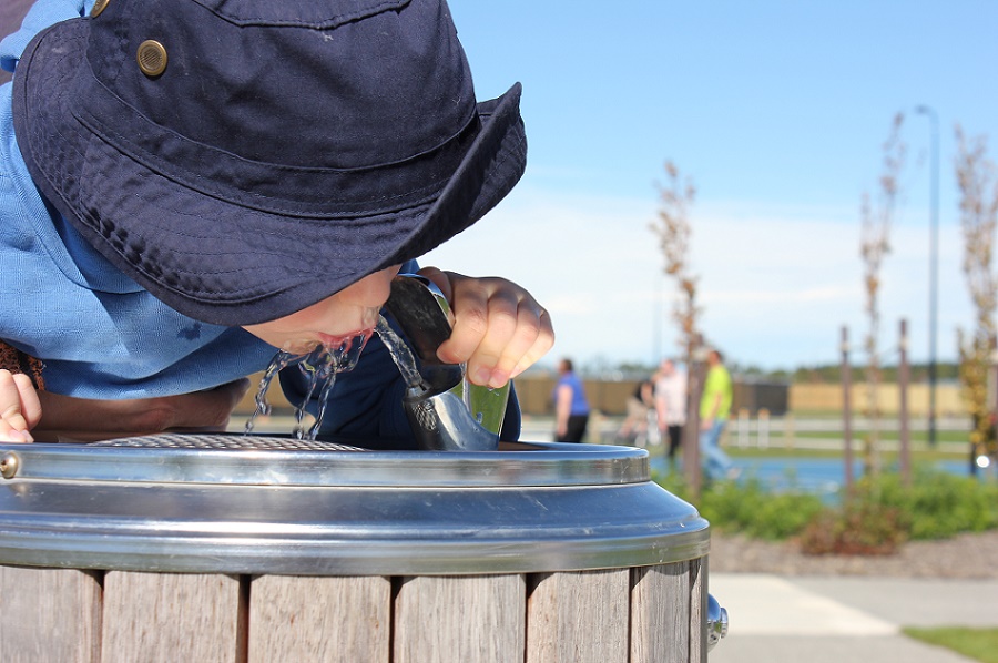 A boy in a blue sun hat drinks from a drinking fountain
