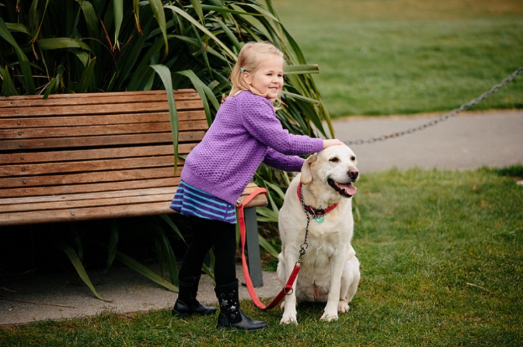Young girl in a purple jersey and black trousers stands in a park with her hands on the head of a blonde labrador retreiver