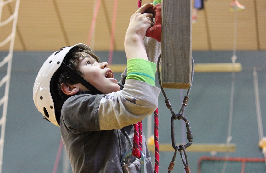 Black haired boy in white climbing helmet and grey t-shirt holding a red climbing hold and looking up for next hold