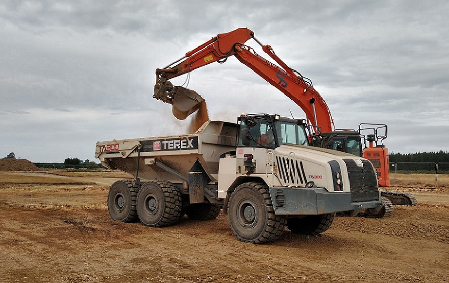 A digger drops a scoop of dirt into the back of a dump track. Both are on a patch of bare brown earth