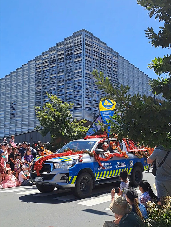 Blue and yellow emergency car decorated in red tinsel and children waving in the background