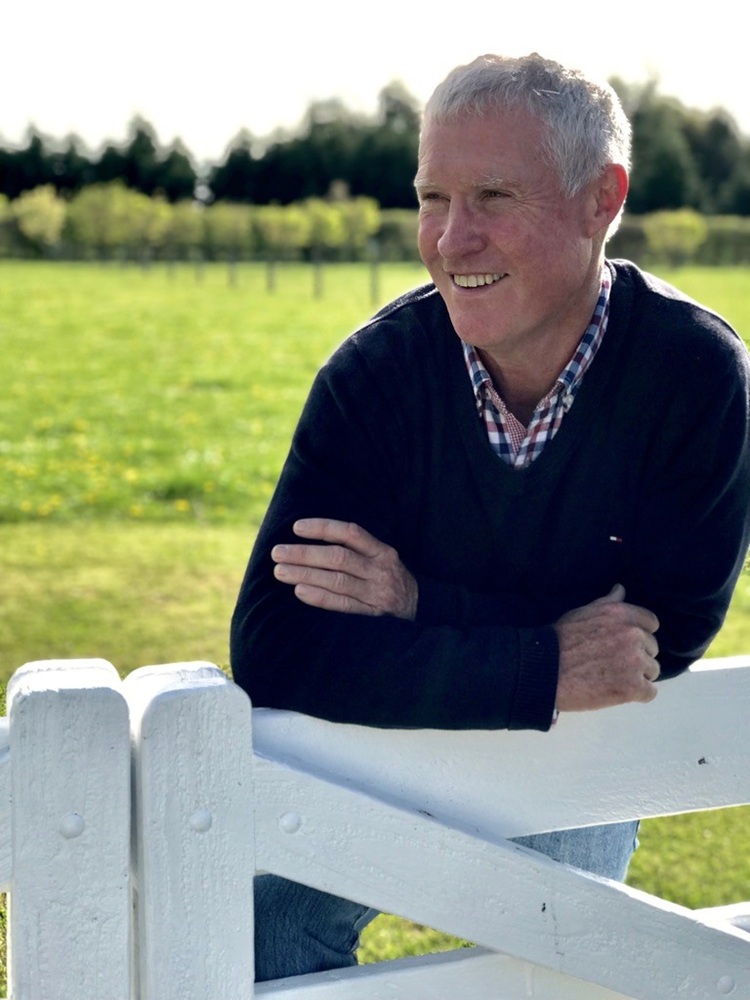 Portrait photo of Bryan Jamison leaning against a  white fence with grass and grape vines behind
