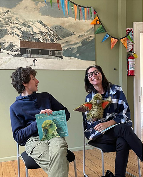 Two people sitting on chairs, the man with a book in hand and the woman with a kea puppet