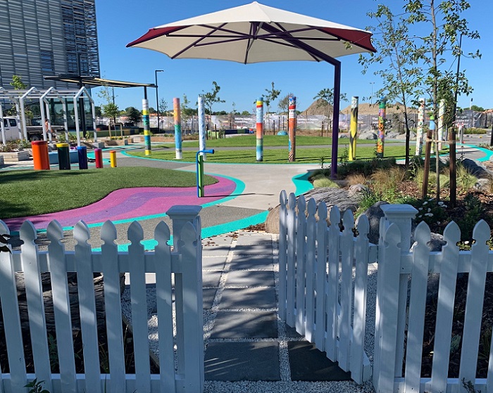 a fence and open gate with the sensory experience behind, looking towards colourful paths and poles
